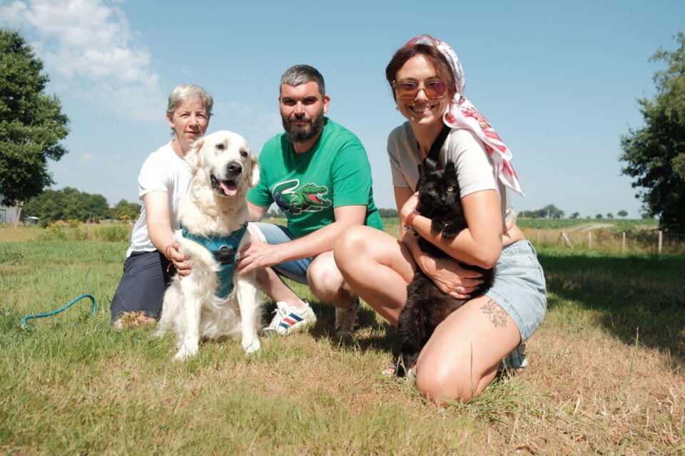 Trois membres du collectif accroupis dans un pré : deux femmes et un homme, avec un grand chien clair et un chat noir dans les bras.